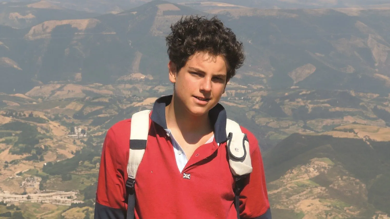 St. Carlo Acutis in a red polo shirt with a backpack, mountains behind him