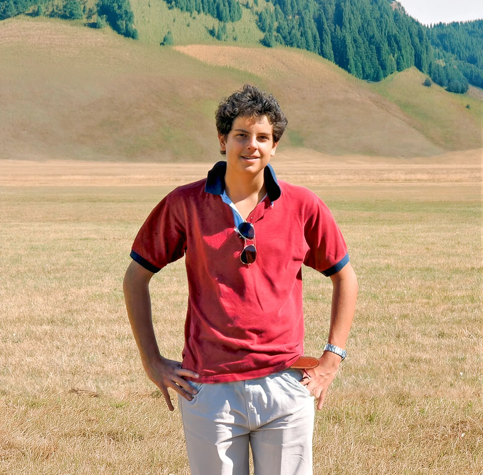 St. Carlo Acutis in a red polo shirt, standing in a field below green hills